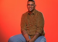 A lightly melanated man with a smile in his face while sitting down on a stool with an orange background 